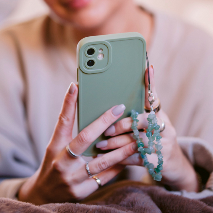 A person with manicured nails holds a smartphone encased in the NAKD Phone Case - Sage by Coconut Lane, featuring an attached beaded charm. The individual is wearing a beige top and various rings. The background is softly blurred, creating a cozy atmosphere.