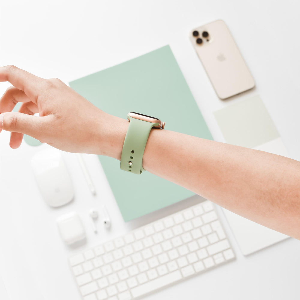 A person wearing a Coconut Lane NAKD Apple Watch Strap in Sage Green on their wrist. The background shows a tiered workstation with items including a light green notebook, a smartphone, a computer mouse, wireless earbuds, a keyboard, a notepad, and color swatches on a white surface.