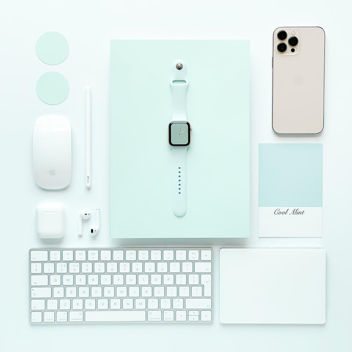 A flat lay of tech gadgets and accessories on a light mint background features an iPhone, Apple Watch with a sleek NAKD Apple Watch Strap in Cool Mint by Coconut Lane, iPad, keyboard, mouse, AirPods, stylus, charger, and a mint-colored card labeled "Cool Mint." The devices are neatly arranged in a grid pattern.