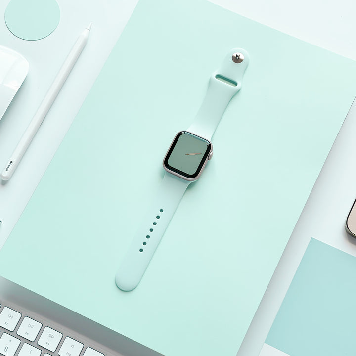 A NAKD Apple Watch Strap in Cool Mint by Coconut Lane, featuring a silicone material and a square screen, lies flat on a matching mint green surface. Surrounding it are various desk items, including a white pencil, part of a white keyboard, and part of a smartphone, all arranged neatly in minimalist style. Part of the NAKD Collection.