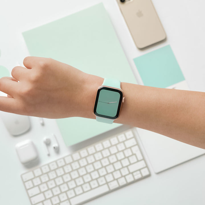 A person's wrist wearing an Apple Watch with the NAKD Apple Watch Strap in Cool Mint from Coconut Lane, positioned over a white desk adorned with various items: a keyboard, earphones, an iPhone, a computer mouse, and neatly arranged papers, all incorporating a white and mint green color scheme.