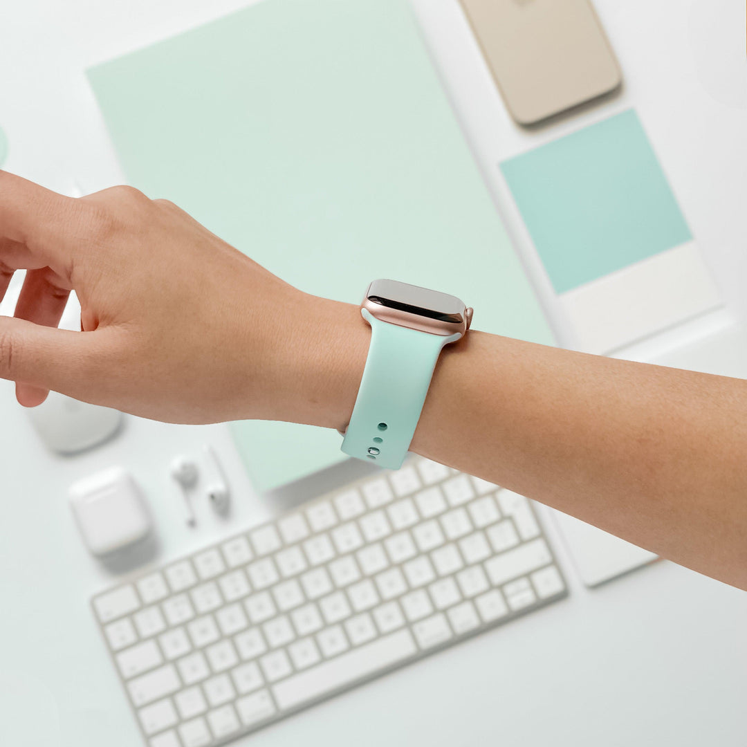 A person's wrist is adorned with a NAKD Apple Watch Strap - Cool Mint by Coconut Lane. The background features minimalistic, color-coordinated items like a keyboard, earphones, and various flat objects in shades of mint green and beige. The overall aesthetic is clean and modern, fitting perfectly with the NAKD Collection.