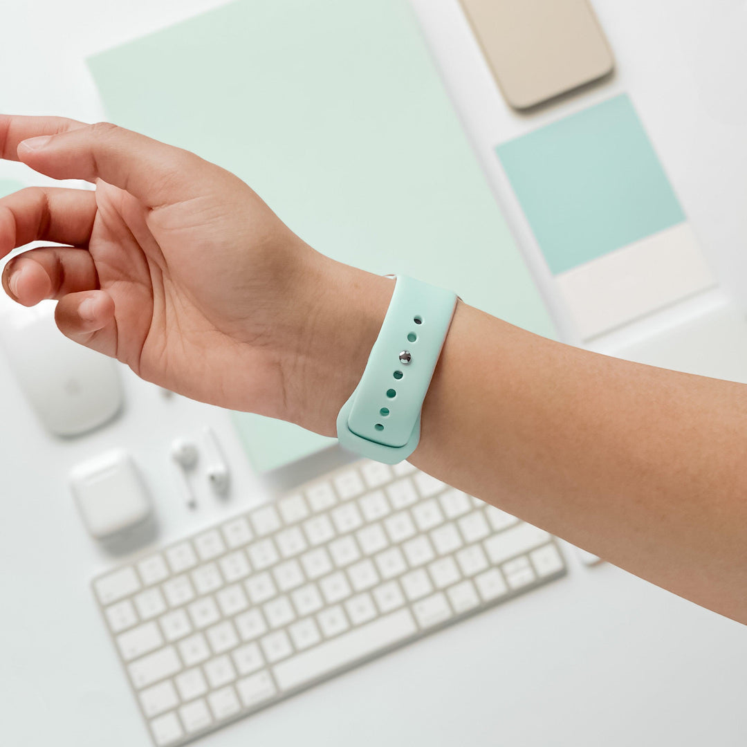 A person's wrist wearing a Cool Mint silicone strap from Coconut Lane's NAKD Apple Watch Strap collection, with a minimalistic workspace background including a white keyboard, mouse, earphones, smartphone, and various stationery items, all in pastel colors.