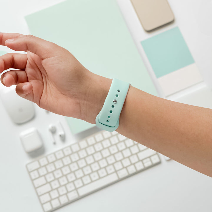 A person's wrist wearing a Cool Mint silicone strap from Coconut Lane's NAKD Apple Watch Strap collection, with a minimalistic workspace background including a white keyboard, mouse, earphones, smartphone, and various stationery items, all in pastel colors.
