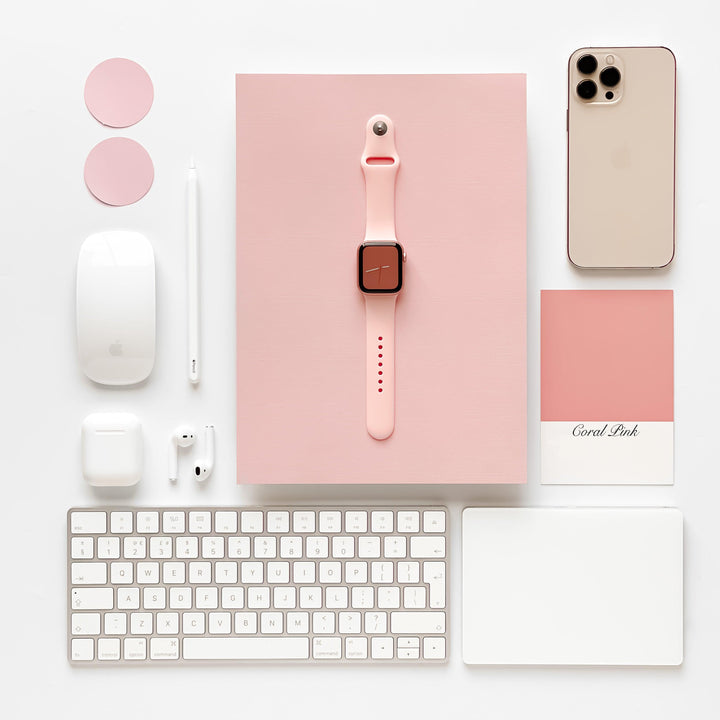 A neatly arranged flat lay featuring a spring-inspired pastels-themed Apple setup including an iPhone, an Apple Watch with a Coconut Lane NAKD Apple Watch Strap - Coral Pink, AirPods, Magic Mouse, Magic Keyboard, an Apple Pencil, a trackpad, and two pink coasters. There is also a pink notebook and a "Coral Pink" color card.