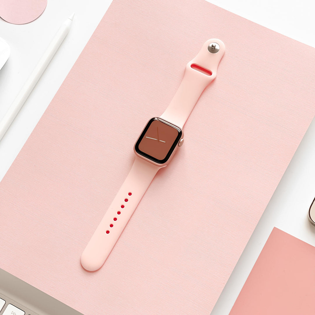 A smartwatch with a NAKD Apple Watch Strap - Coral Pink from Coconut Lane is displayed on a pink surface. The watch face shows a simple, clean clock design. There is a pencil and a paper clip partially visible on the left side of the image, and a smartphone on the bottom right corner.