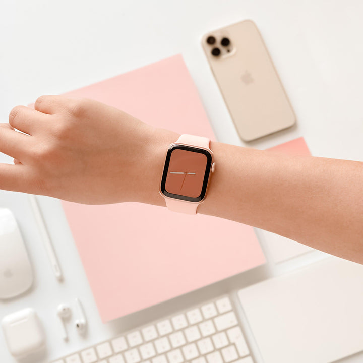 A person's arm with a Coconut Lane NAKD Apple Watch Strap in Coral Pink, featuring a pink face, resting on a white surface. Nearby are a smartphone with three cameras, a computer mouse, a keyboard, earphones, and pastel spring-inspired notepads, all arranged neatly.