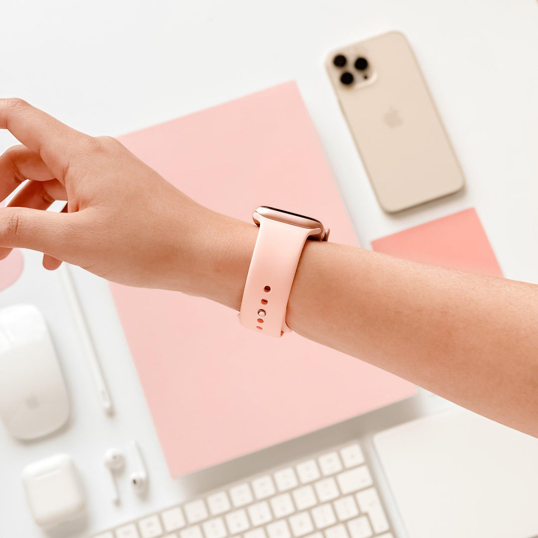 A hand showcasing the NAKD Apple Watch Strap - Coral Pink by Coconut Lane, surrounded by spring-inspired pastel tech accessories—including an iPhone, keyboard, mouse, earphones, and a pink notebook—beautifully arranged on a clean, light surface.