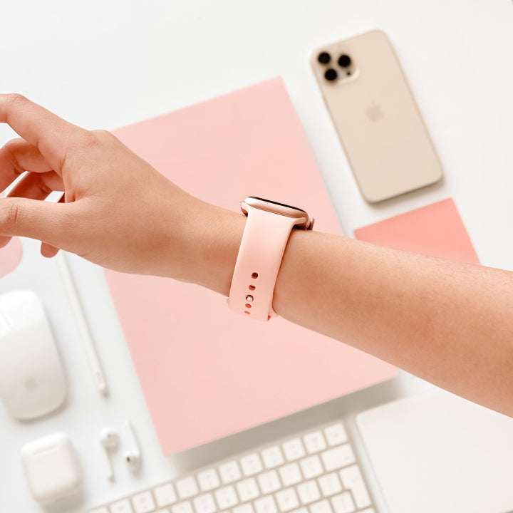 A hand showcasing the NAKD Apple Watch Strap - Coral Pink by Coconut Lane, surrounded by spring-inspired pastel tech accessories—including an iPhone, keyboard, mouse, earphones, and a pink notebook—beautifully arranged on a clean, light surface.