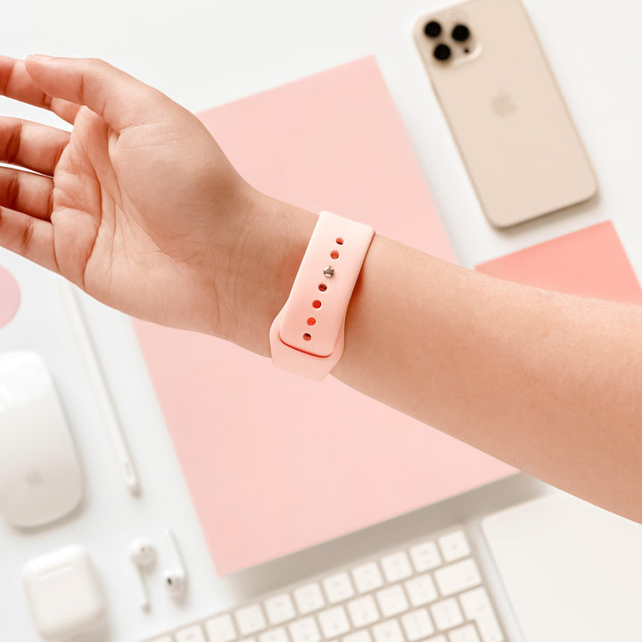 A person is wearing a Coconut Lane NAKD Apple Watch Strap - Coral Pink on their wrist. The background features a beige smartphone, a closed laptop, wireless earbuds in a case, a wireless mouse, a stylus, a keyboard, and pink square papers. The overall color scheme is spring inspired pastels.