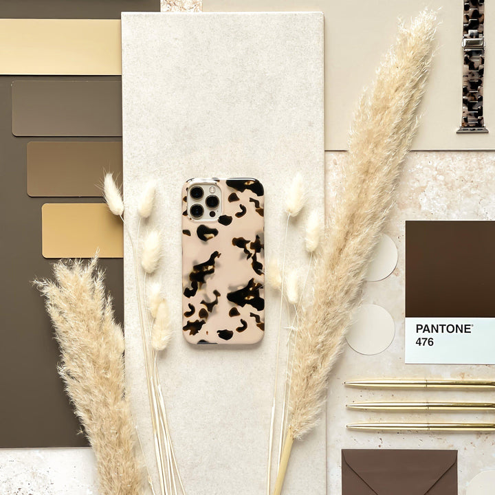 A flat lay photo featuring the Ivory Tort Phone Case from Coconut Lane on a textured beige surface, surrounded by dried pampas grass, color swatches in various shades of brown, and Pantone color cards. Two gold pens and a brown envelope are neatly arranged to the side.