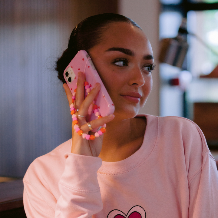 A person with a sleek hairstyle holds a pink phone to their ear, showcasing the Coconut Lane Cute Hearts Phone Case that perfectly matches their pink pearl phone charm. Wearing a light pink sweatshirt adorned with a heart design, they stand out against the blurred background, creating a charming Valentine's Day vibe.